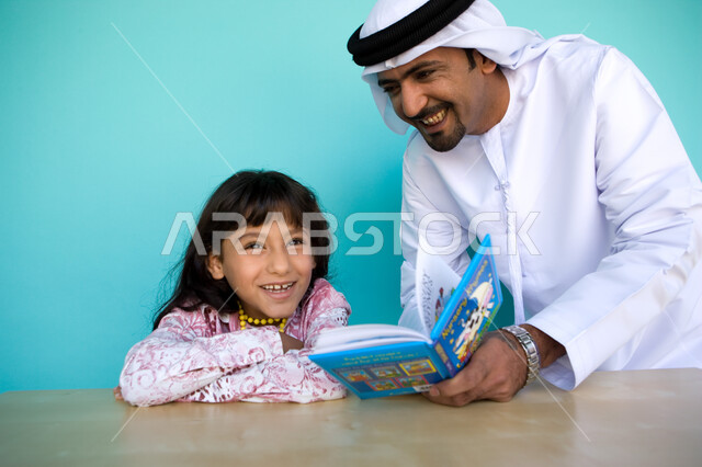 الاستمتاع والضحك مع الأب أثناء المطالعة, تشجيع الأبناء على التعلم والتقف, بورتريه مقرب لرجل عربي خليجي إماراتي يرتدي الغترة والكندورة الإماراتية يقوم بمساعدة ابنته على القراءة, مطالعة القصص التصويرية المسلية, تنمية مهارات القراءة للأطفال, خلفية زرقاء