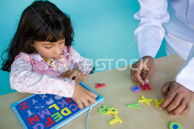 التسلية والترفيه بالأنشطة التعليمية, الاهتمام بتعليم وترفيه الأبناء, التعلم عن طريق الألعاب التثقيفية، التعرف على أشكال الحروف الأجنبية، بورتريه مقرب من الأعلى لفتاة عربية خليجية إماراتية تقوم بحل لغز الأبجدية بمساعدة والدها , خلفية زرقاء