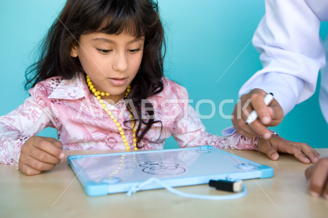 الاهتمام بمواهب الأطفال ورعايتها, دور الأهل في تنمية المهارات الفنية, إيماءات السعادة والاستمتاع بمشاركة الأنشطة الفنية مع الأب، شغف وحب الرسم، بورتريه مقرب لفتاة صغيرة عربية خليجية إماراتية تقوم بالرسم على اللوح بمساعدة والدها، خلفية زرقاء