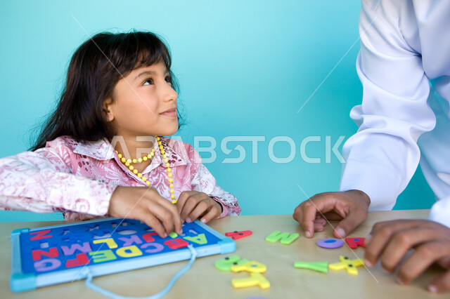 الاهتمام بتعليم وترفيه الأبناء, التسلية والترفيه بالأنشطة التعليمية, التعلم عن طريق الألعاب التثقيفية، التعرف على أشكال الحروف الأجنبية، بورتريه مقرب لفتاة عربية خليجية إماراتية تقوم بحل لغز الأبجدية بمساعدة والدها وتنظر اليه بإيماءات الحب , خلفية زرقاء