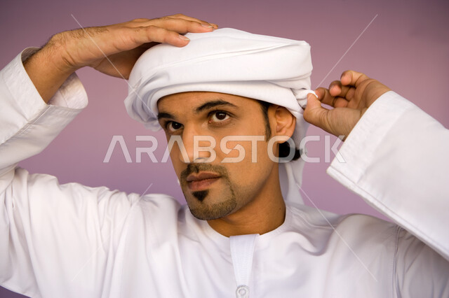 ارتداء الكندورة الاماراتية والعمامة, النظر إلى شيء ما بإيماءات التركيز, الاهتمام بالمظهر الخارجي, بورتريه مقرب لشاب عربي خليجي إماراتي يرتدي الزي التقليدي الإماراتي يقوم بلف العمامة, خلفية ملونة