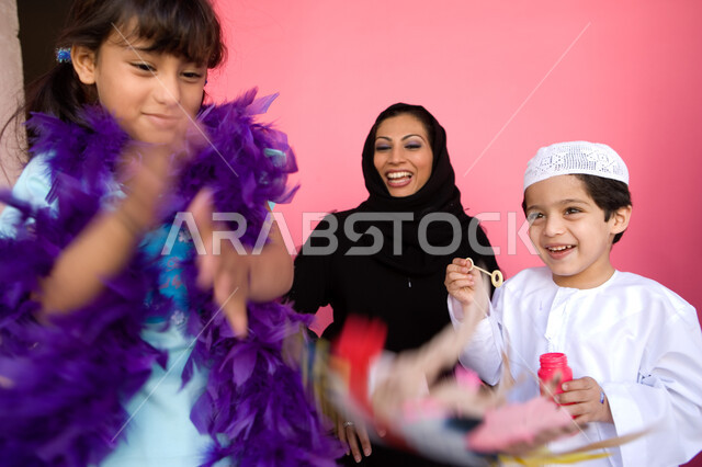 اللعب بالفقاعات المائية بإيماءات السعادة والفرح, قضاء الأوقات مع الأسرة, بورتريه مقرب لعائلة عربية خليجية إماراتية تستمتع باللعب معا, تنمية مهارات الأطفال الحركية, مفهوم الأسرة السعيدة, زرع روح المحبة بين الأبناء, خلفية وردية