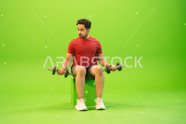 القوة والتحدي والتحفيز لأسلوب حياة صحي، نشاط بدني للمحافظة على الصحة واللياقة الجسدية، رفع الأوزان الحديدية الثقيلة، تقوية عضلات الجسم وبناؤها، تمارين رفع الأثقال، بورتريه لشاب عربي خليجي سعودي يمارس التمارين الرياضية مع الدمبلز، خلفية خضراء، كروما.