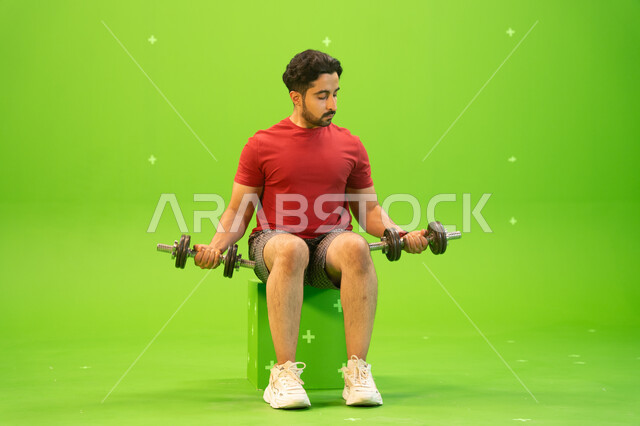 القوة والتحدي والتحفيز لأسلوب حياة صحي، نشاط بدني للمحافظة على الصحة واللياقة الجسدية، رفع الأوزان الحديدية الثقيلة، تقوية عضلات الجسم وبناؤها، تمارين رفع الأثقال، بورتريه لشاب عربي خليجي سعودي يمارس التمارين الرياضية مع الدمبلز، خلفية خضراء، كروما.