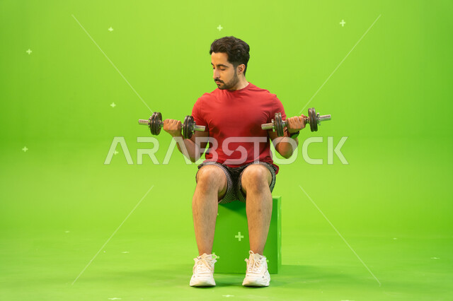 نشاط بدني للمحافظة على الصحة واللياقة الجسدية، رفع الأوزان الحديدية الثقيلة، تقوية عضلات الجسم وبناؤها، تمارين رفع الأثقال، القوة والتحدي والتحفيز لأسلوب حياة صحي، بورتريه لشاب عربي خليجي سعودي يمارس التمارين الرياضية مع الدمبلز، خلفية خضراء، كروما.
