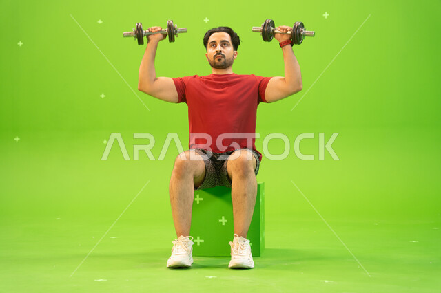 تقوية عضلات الجسم وبناؤها، تمارين رفع الأثقال، القوة والتحدي والتحفيز لأسلوب حياة صحي، بورتريه لشاب عربي خليجي سعودي يمارس التمارين الرياضية مع الدمبلز، نشاط بدني للمحافظة على الصحة واللياقة الجسدية، رفع الأوزان الحديدية الثقيلة، خلفية خضراء، كروما.