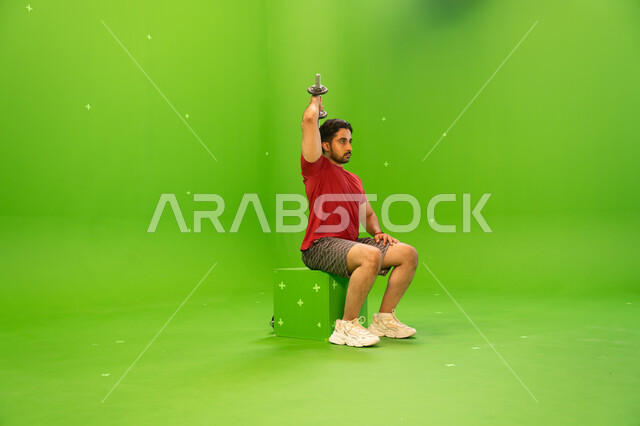 نشاط بدني للمحافظة على الصحة واللياقة الجسدية، رفع الأوزان الحديدية الثقيلة، تقوية عضلات الجسم وبناؤها، تمارين رفع الأثقال، القوة والتحدي والتحفيز لأسلوب حياة صحي، بورتريه لشاب عربي خليجي سعودي يمارس التمارين الرياضية مع الدمبلز، خلفية خضراء، كروما.