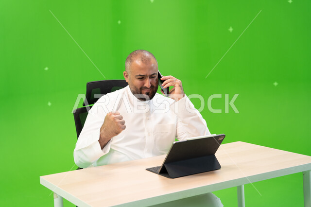 الربح في التجارة الالكترونية والبورصة، إيماءات النصر والفوز، الفرح بالإنجاز وتحقيق الأهداف, بورتريه مقرب لموظف سعودي خليجي يرتدي الثوب السعودي التقليدي يقوم بإجراء مكالمة هاتفية عبر الهاتف المحمول داخل مكتبه, النظر إلى جهاز التابليت, خلفية خضراء, كروما.