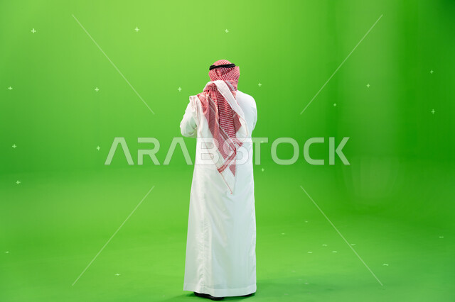 النظر إلى شيء ما، صورة الجسم بالطول الكامل، الوقوف باستقامة، بورتريه من الخلف لرجل عربي خليجي سعودي يرتدي الشماغ والثوب التقليدي السعودي، الاهتمام بالمظهر الخارجي، خلفية خضراء، كروما.