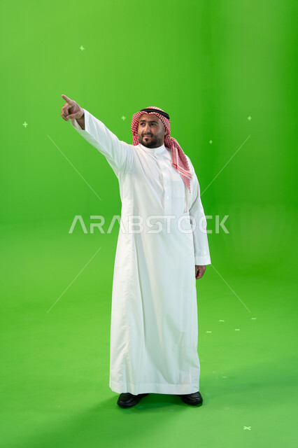 الإشارة بإصبع السبابة إلى شيء ما، صورة الجسم بالطول الكامل، الوقوف باستقامة، مفهوم لغة الجسد والحركات والإشارات، بورتريه لرجل عربي خليجي سعودي يرتدي الشماغ والثوب التقليدي السعودي، خلفية خضراء، كروما.