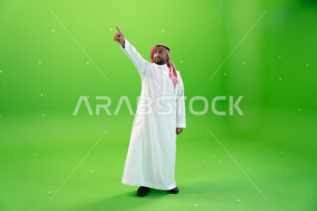 الإشارة بإصبع السبابة إلى شيء ما، صورة الجسم بالطول الكامل، الوقوف باستقامة، مفهوم لغة الجسد والحركات والإشارات، بورتريه لرجل عربي خليجي سعودي يرتدي الشماغ والثوب التقليدي السعودي، خلفية خضراء، كروما.
