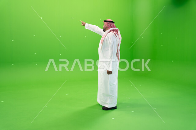 صورة الجسم بالطول الكامل، الوقوف باستقامة، مفهوم لغة الجسد والحركات والإشارات، بورتريه من الجانب لرجل عربي خليجي سعودي يرتدي الشماغ والثوب التقليدي السعودي يقوم بالإشارة بإصبع السبابة إلى شيء ما، خلفية خضراء، كروما.