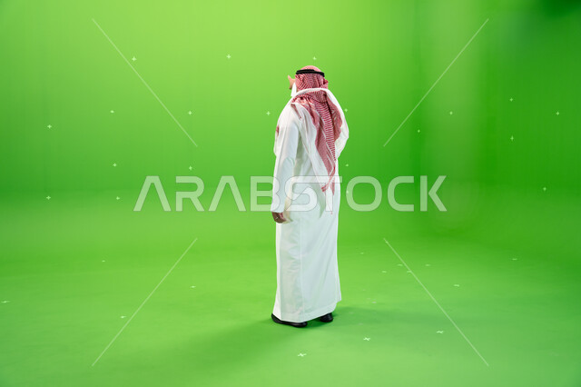 مفهوم لغة الجسد والحركات والإشارات، بورتريه من الخلف لرجل عربي خليجي سعودي يرتدي الشماغ والثوب التقليدي السعودي يقوم بالإشارة بإصبع السبابة إلى شيء ما، صورة الجسم بالطول الكامل، الوقوف باستقامة، خلفية خضراء، كروما.