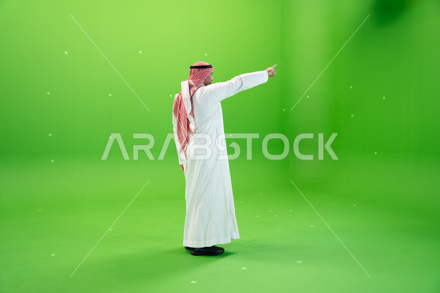 صورة الجسم بالطول الكامل، الوقوف باستقامة، مفهوم لغة الجسد والحركات والإشارات، بورتريه من الجانب لرجل عربي خليجي سعودي يرتدي الشماغ والثوب التقليدي السعودي يقوم بالإشارة بإصبع السبابة إلى شيء ما، خلفية خضراء، كروما.