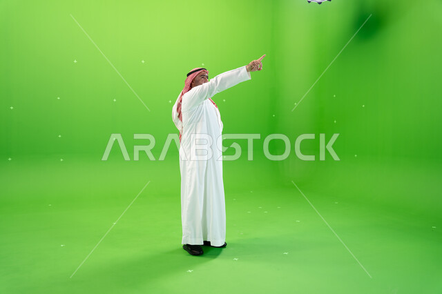 صورة الجسم بالطول الكامل، الوقوف باستقامة، مفهوم لغة الجسد والحركات والإشارات، بورتريه من الجانب لرجل عربي خليجي سعودي يرتدي الشماغ والثوب التقليدي السعودي يقوم بالإشارة بإصبع السبابة إلى شيء ما، خلفية خضراء، كروما.