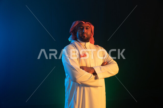 شاب وسيم ينظر الى الكاميرا بإيماءات ايدي متقاطعه ،شخصيه سعوديه بارزه ،الثقه والاعتزاز بالنفس ، بورتريه لرجل عربي خليجي سعودي مبتسم يرتدي الثوب والشماغ التقليدي يقف باستقامة، مفهوم الشياكه والاهتمام بالمظهر الخارجي، خلفية سوداء مع إضاءة ملونه