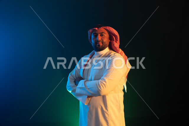 شخصيه سعوديه بارزه، النظر إلى الكاميرا بإيماءات أيدي متقاطعه والثقه والاعتزاز بالنفس، مفهوم الاهتمام بالمظهر الخارجي والشياكة ، بورتريه  لرجل عربي خليجي سعودي مبتسم يرتدي الثوب والشماغ التقليدي ، خلفية سوداء مع إضاءة ملونة