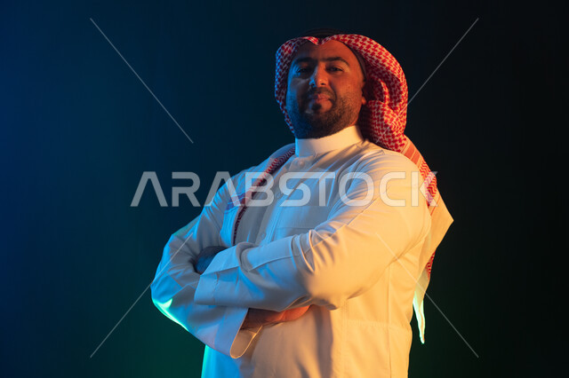  النظر إلى الكاميرا بإيماءات أيدي متقاطعه والثقه والاعتزاز بالنفس، شخصيه سعوديه مهمة ، مفهوم الاهتمام بالمظهر الخارجي والشياكة ، بورتريه مقرب من الجانب لرجل عربي خليجي سعودي مبتسم يرتدي الثوب والشماغ التقليدي ، خلفية سوداء مع إضاءة ملونة