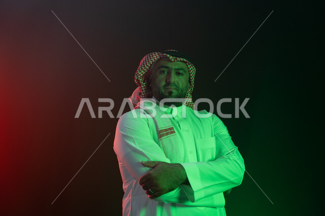 A prominent Saudi personality, standing with crossed hand gestures, confidence and self-esteem, contemplating and looking at the camera, the concept of caring for external appearance and elegance, a close-up portrait of a smiling Saudi Gulf Arab man weari