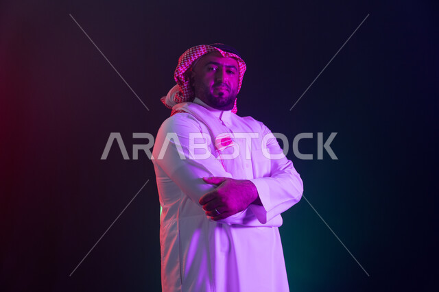 A prominent Saudi personality, standing with crossed hand gestures, confidence and self-esteem, contemplating and looking at the camera, the concept of caring for external appearance and elegance, a close-up portrait of a smiling Saudi Gulf Arab man weari