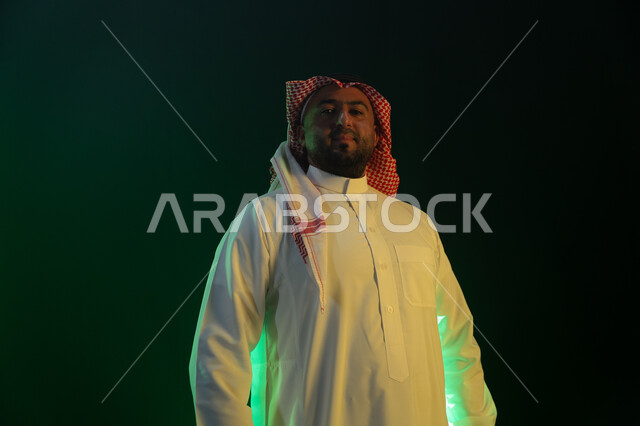 النظر إلى الكاميرا بإيماءات السعاده، الثقه بالنفس والاعتزاز، مفهوم الاهتمام بالمظهر الخارجي والشياكة ،شخصيه سعوديه بارزه ، بورتريه مقرب لرجل عربي خليجي سعودي مبتسم يرتدي الثوب والشماغ التقليدي، خلفية سوداء مع إضاءة ملونه