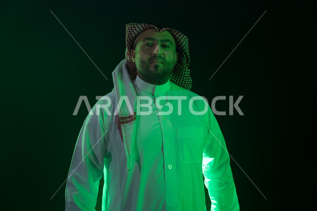 حركات الثقه بالنفس والاعتزاز، مفهوم الاهتمام بالمظهر الخارجي والشياكة، شاب وسيم ينظر الى الكاميرا ،شخصيه سعوديه بارزه ، بورتريه مقرب لرجل عربي خليجي سعودي مبتسم يرتدي الثوب والشماغ التقليدي، خلفية سوداء مع إضاءة ملونه