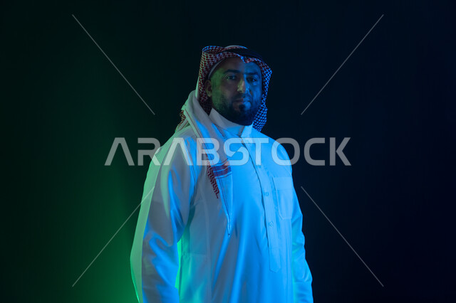 حركات الثقه بالنفس والاعتزاز، مفهوم الاهتمام بالمظهر الخارجي والشياكة، شاب وسيم ينظر الى الكاميرا ،شخصيه سعوديه بارزه ، بورتريه مقرب لرجل عربي خليجي سعودي مبتسم يرتدي الثوب والشماغ التقليدي، خلفية سوداء مع إضاءة ملونه