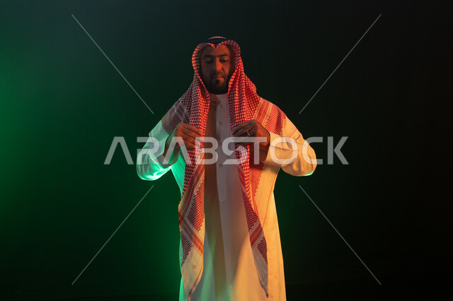 إيماءات تعديل اللباس ،مفهوم الاهتمام بالمظهر الخارجي والشياكة ،الثقه والاعتزاز بالنفس،شخصيه سعوديه بارزه ، بورتريه لرجل عربي خليجي سعودي مبتسم يرتدي الثوب والشماغ التقليدي ، خلفية سوداء مع إضاءة ملونه