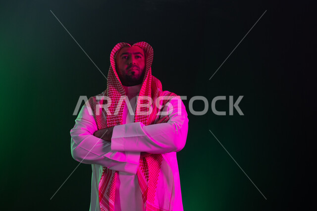 الوقوف بإيماءات أيدي متقاطعه والثقه والاعتزاز بالنفس، التأمل والنظر إلى شيء ما، مفهوم الاهتمام بالمظهر الخارجي والشياكة ،شخصيه سعوديه بارزه ، بورتريه مقرب لرجل عربي خليجي سعودي مبتسم يرتدي الثوب والشماغ التقليدي ، خلفية سوداء مع إضاءة ملونه