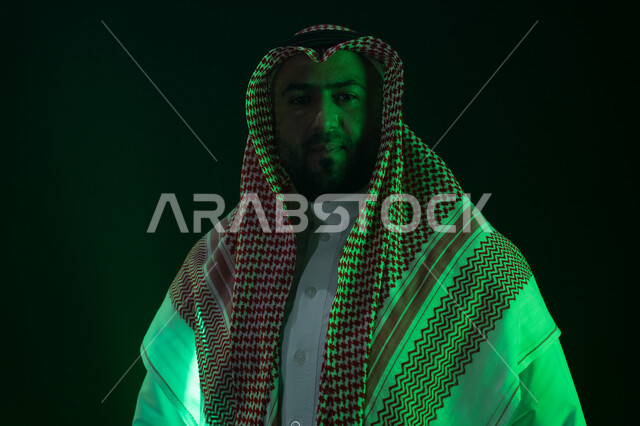 مفهوم الاهتمام بالمظهر الخارجي والشياكة، شاب وسيم ينظر الى الكاميرا ،شخصيه سعوديه بارزه ،ايماءات الثقه بالنفس والاعتزاز، بورتريه مقرب لرجل عربي خليجي سعودي مبتسم يرتدي الثوب والشماغ التقليدي يقف باستقامة، خلفية سوداء مع إضاءة ملونه