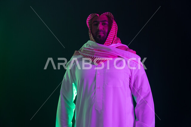 شخصيه سعوديه بارزه ،ايماءات الثقه بالنفس والاعتزاز، بورتريه مقرب لرجل عربي خليجي سعودي مبتسم يرتدي الثوب والشماغ التقليدي يقف باستقامة، النظر إلى الكاميرا، مفهوم الشياكه والاهتمام بالمظهر الخارجي، خلفية سوداء مع إضاءة ملونه
