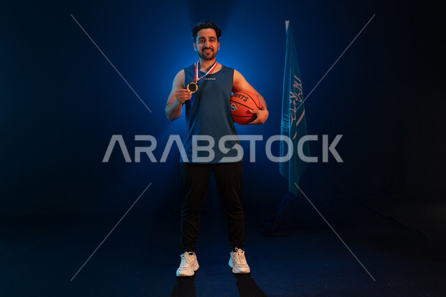 Standing with gestures of happiness and joy over victory, a Saudi Arabian Gulf player wearing a sports uniform and a gold medal holding the ball standing next to the Saudi flag, commemorating National Day September 23, Saudi National Basketball Team, Flag