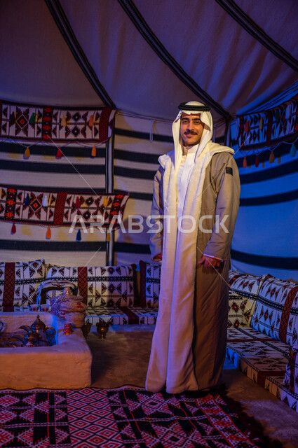 إيماءات وجه تدل على السرور، رحلة وكشتة بر في الأجواء الشتوية في الصحراء، خيمة بدوية شعبية تراثية في السعودية، الأجواء الهادئة في أماكن التخييم، رجل عربي خليجي سعودي مبتسم يرتدي الفروة والغترة يقف داخل بيت شعر ينظر إلى الكاميرا، صورة الجسم بالطول الكامل