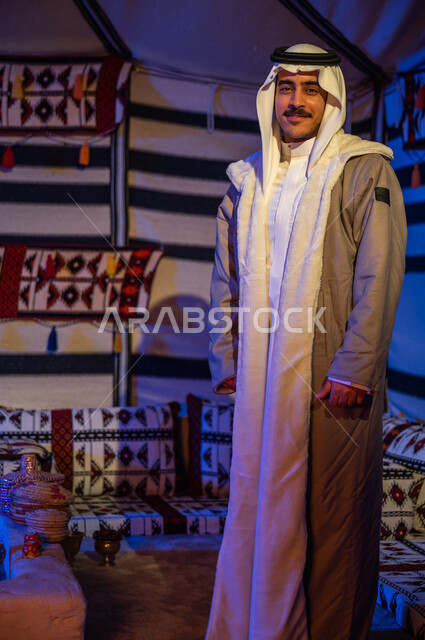 رحلة وكشتة بر في الأجواء الشتوية في الصحراء، خيمة بدوية شعبية تراثية في السعودية ، الأجواء الهادئة في أماكن التخييم، رجل عربي خليجي سعودي مبتسم يرتدي الفروة والغترة التقليدية يقف في داخل بيت شعر ينظر إلى الكاميرا، إيماءات وجه تدل على السرور، صورة الجسم بالطول الكامل