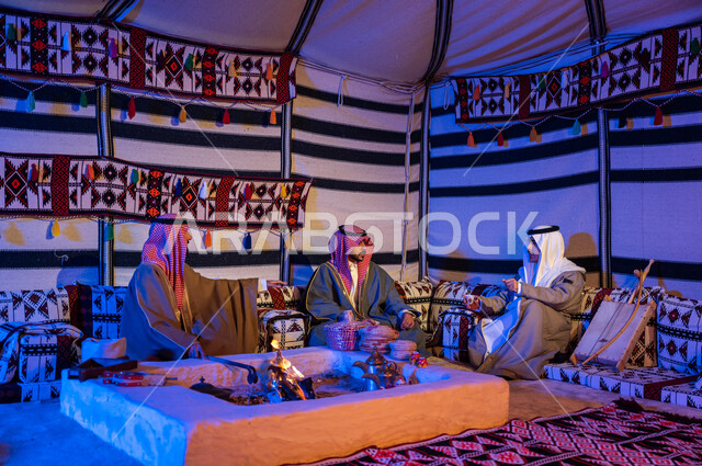 كشتة بر شبابية داخل خيمة تراثية، أصدقاء عرب خليجيون سعوديون يرتدون الازياء الوطنية يتبادلون الاحاديث في جلسات شعبية، عادات وتقاليد سعودية في تقديم واعداد القهوة العربية وكرم الضيافه، تحريك الجمر في الموقد الطيني باستخدام الملقط