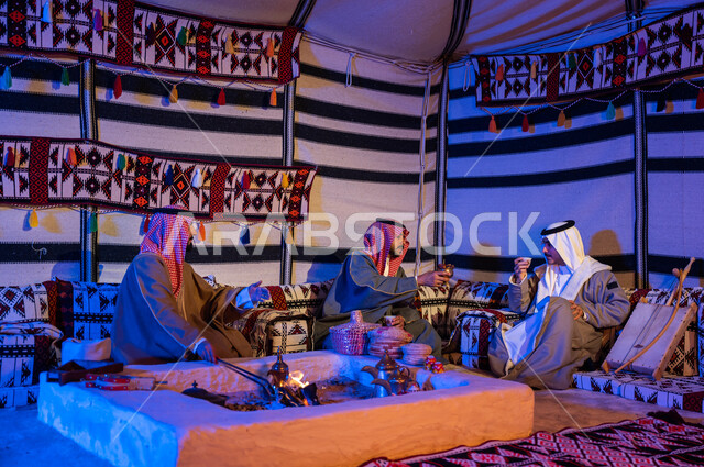 أصدقاء عرب خليجيون سعوديون يرتدون الازياء الوطنية يتبادلون الاحاديث في جلسات شعبية، عادات وتقاليد سعودية في تقديم واعداد القهوة العربية وكرم الضيافه، تحريك الجمر في الموقد الطيني باستخدام الملقط، كشتة بر شبابية داخل خيمة تراثية