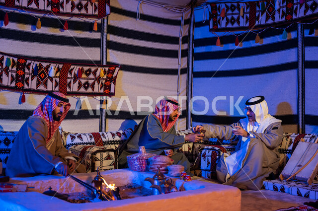 أصدقاء عرب خليجيون سعوديون يرتدون الازياء الوطنية يتبادلون الاحاديث في جلسات شعبية، عادات وتقاليد سعودية في تقديم واعداد القهوة العربية وكرم الضيافه، تحريك الجمر في الموقد الطيني باستخدام الملقط، كشتة بر شبابية داخل خيمة تراثية
