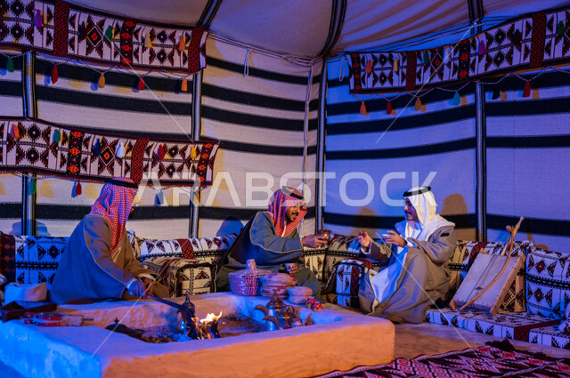 Saudi Gulf Arab friends wearing national costumes exchange conversations in popular gatherings, Saudi customs and traditions in serving and preparing Arabic coffee and generous hospitality, stirring coals in the clay stove using tongs, youthful wheat kashta inside a heritage tent.