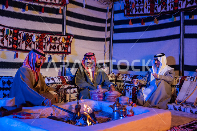 أصدقاء عرب خليجيون سعوديون يرتدون الازياء الوطنية يتبادلون الاحاديث في جلسات شعبية، عادات وتقاليد سعودية في تقديم واعداد القهوة العربية وكرم الضيافه، تحريك الجمر في الموقد الطيني باستخدام الملقط، كشتة بر شبابية داخل خيمة تراثية