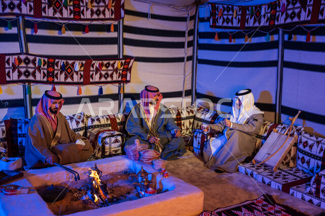Saudi Gulf Arab friends wearing national costumes exchange conversations in popular gatherings, Saudi customs and traditions in serving and preparing Arabic coffee and generous hospitality, stirring coals in the clay stove using tongs, youthful wheat kashta inside a heritage tent.