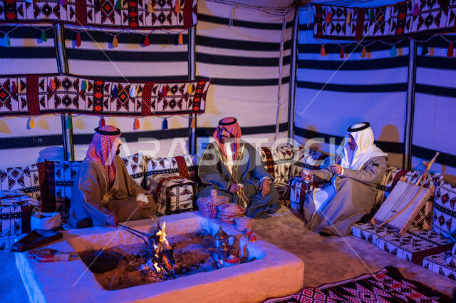 عادات وتقاليد سعودية في تقديم واعداد القهوة العربية وكرم الضيافه، تحريك الجمر في الموقد الطيني باستخدام الملقط، كشتة بر شبابية داخل خيمة تراثية، ارتداء الازياء الوطنية، أصدقاء عرب خليجيون سعوديون يرتديون الشماغ والبشت يتبادلون الاحاديث في جلسات شعبية