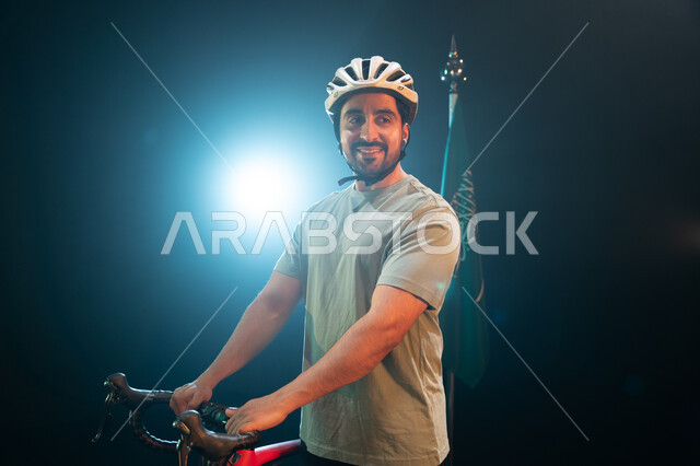 نشاط بدني للمحافظه على الصحه, الوقوف بجانب البيرق والنظر إلى الكاميرا، بورتريه لرجل سعودي يرتدي زي الرياضة الخاص بسباق الدراجات الهوائية ،ممارسة هواية ركوب الدراجات، يوم العلم 11 مارس، ذكرى اليوم الوطني السعودي 23 سبتمبر، خلفية سوداء مع اضاءة بيضاء