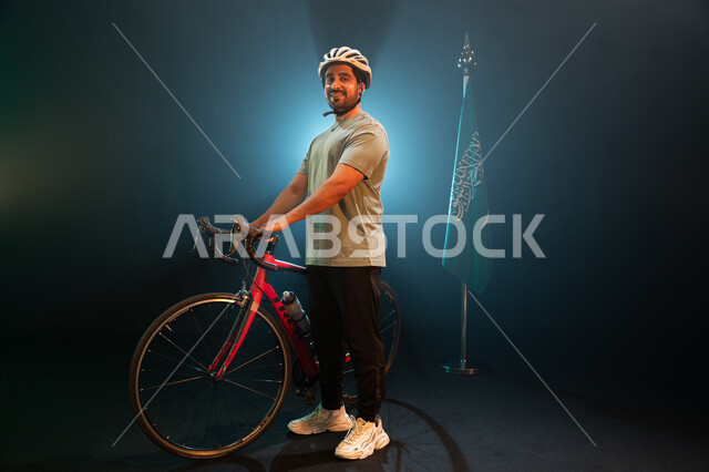 يوم العلم 11 مارس، هواية ركوب الدراجات، الوقوف بجانب البيرق والنظر إلى الكاميرا بإيماءات السعادة، بورتريه لرجل سعودي يرتدي زي الرياضة الخاص بسباق الدراجات الهوائية ،ذكرى اليوم الوطني السعودي 23 سبتمبر، صورة الجسم بالطول الكامل، خلفية سوداء مع اضاءة بيضاء