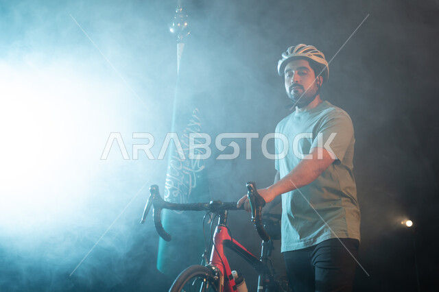 نشاط بدني للمحافظه على الصحه الجسديه, بورتريه لرجل سعودي يرتدي زي الرياضة الخاص بسباق الدراجات الهوائية يقف بجانب البيرق وينظر إلى الكاميرا،ممارسة هواية ركوب الدراجات، يوم العلم 11 مارس، ذكرى اليوم الوطني السعودي 23 سبتمبر، خلفية سوداء مع اضاءة بيضاء