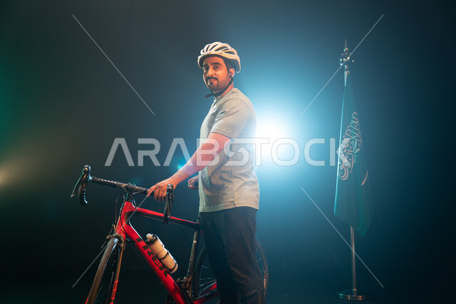 الوقوف بجانب البيرق والنظر إلى الكاميرا، بورتريه لرجل سعودي يرتدي زي الرياضة الخاص بسباق الدراجات الهوائية ،ممارسة هواية ركوب الدراجات، يوم العلم 11 مارس، نشاط بدني للمحافظه على اللياقه الجسديه, ذكرى اليوم الوطني السعودي 23 سبتمبر، خلفية سوداء مع اضاءة بي