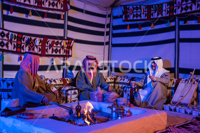 عادات وتقاليد سعودية في تقديم واعداد القهوة العربية وكرم الضيافه، تحريك الجمر في الموقد الطيني باستخدام الملقط، كشتة بر شبابية داخل خيمة تراثية، ارتداء الازياء الوطنية، أصدقاء عرب خليجيون سعوديون يرتديون الشماغ والبشت يتبادلون الاحاديث في جلسات شعبية
