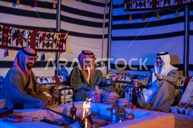 عادات وتقاليد سعودية في تقديم واعداد القهوة العربية وكرم الضيافه، تحريك الجمر في الموقد الطيني باستخدام الملقط، كشتة بر شبابية داخل خيمة تراثية، ارتداء الازياء الوطنية، أصدقاء عرب خليجيون سعوديون يرتديون الشماغ والبشت يتبادلون الاحاديث في جلسات شعبية