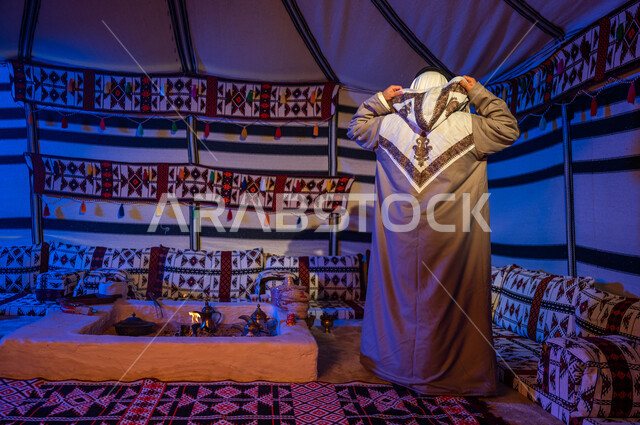 كشتة ورحلة بر في الأجواء الشتوية في الصحراء، خيمة بدوية شعبية تراثية في السعودية، الأجواء الهادئة في أماكن التخييم، صورة من الخلف لرجل عربي خليجي سعودي يقوم بارتداء الفروة والغترة التقليدية يقف في داخل بيت شعر، صورة الجسم بالطول الكامل