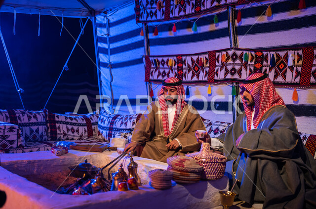 ارتداء الازياء الوطنية، كشتة بر شبابية داخل خيمة تراثية، عادات وتقاليد سعودية في تقديم واعداد القهوة العربية وكرم الضيافه، تحريك الجمر في الموقد الطيني باستخدام الملقط، صديقان عربيان خليجيان سعوديان يرتديان الشماغ والبشت يتبادلان الاحاديث في جلسات شعبية