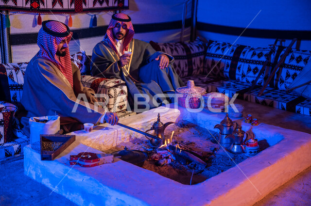 ارتداء الازياء الوطنية، كشتة بر شبابية داخل خيمة تراثية، عادات وتقاليد سعودية في تقديم واعداد القهوة العربية وكرم الضيافه، تحريك الجمر في الموقد الطيني باستخدام الملقط، صديقان عربيان خليجيان سعوديان يرتديان الشماغ والبشت يتبادلان الاحاديث في جلسات شعبية